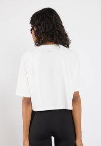 Afskåret hvid T-shirt med korte ærmer, lavet af blødt bomuldsstof. Har et diskret logo på bagsiden af halsudskæringen. Sammen med sorte leggings.
