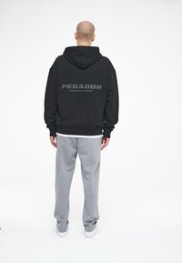 Schwarze Kapuzenjacke mit dem Schriftzug "PEGADOR" auf dem Rücken, kombiniert mit hellgrauen Hosen. Der Stoff wirkt weich und hat eine lockere Passform.