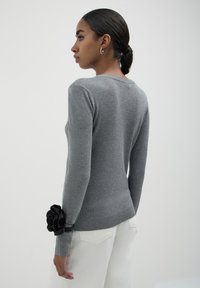 Grauer langärmeliger Pullover mit rundem Ausschnitt und Rippbund, der ein großes schwarzes Blumenmotiv am linken Handgelenk aufweist.