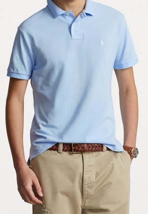 Polo bleu clair en tissu texturé, avec un col, des manches courtes et un petit logo sur la poitrine. Porté avec un pantalon kaki.