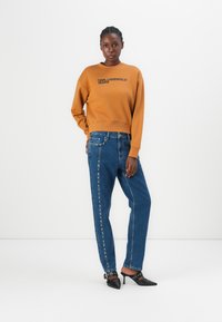 Bruine sweatshirt met "KARL LAGERFELD JEANS" in het zwart gedrukt; blauwe jeans met zijdelingse studs; zwarte puntige hakken.