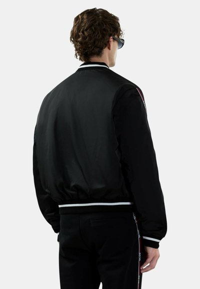 Mężczyzna w czarnej kurtce bomberce z mankietami w białe paski oraz czarnych spodniach z bocznymi paskami z logo, stoi tyłem na szarym tle.