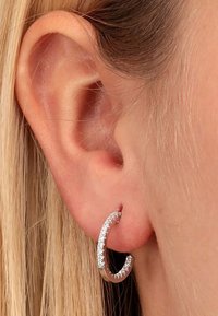 Sølv hoops ørering med en buet form og glitrende udsmykninger, båret i et piercet øre, mod strittende blond hår.