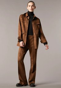 Giacca in finta pelle scamosciata marrone con tasche sul petto, abbinata a pantaloni a gamba larga coordinati e un dolcevita nero, con scarpe nere chunky.