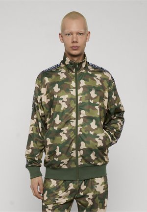 Ung person med kort blondt hår iført en grøn og brun camouflage lynlåstrøje og matchende bukser, hænderne i lommerne.