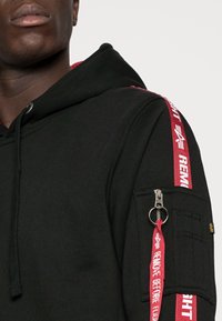Sudadera negra con cinta roja a los lados que dice "RETIRAR ANTES DEL VUELO", bolsillo con cremallera en la manga, capucha con cordón y textura suave.