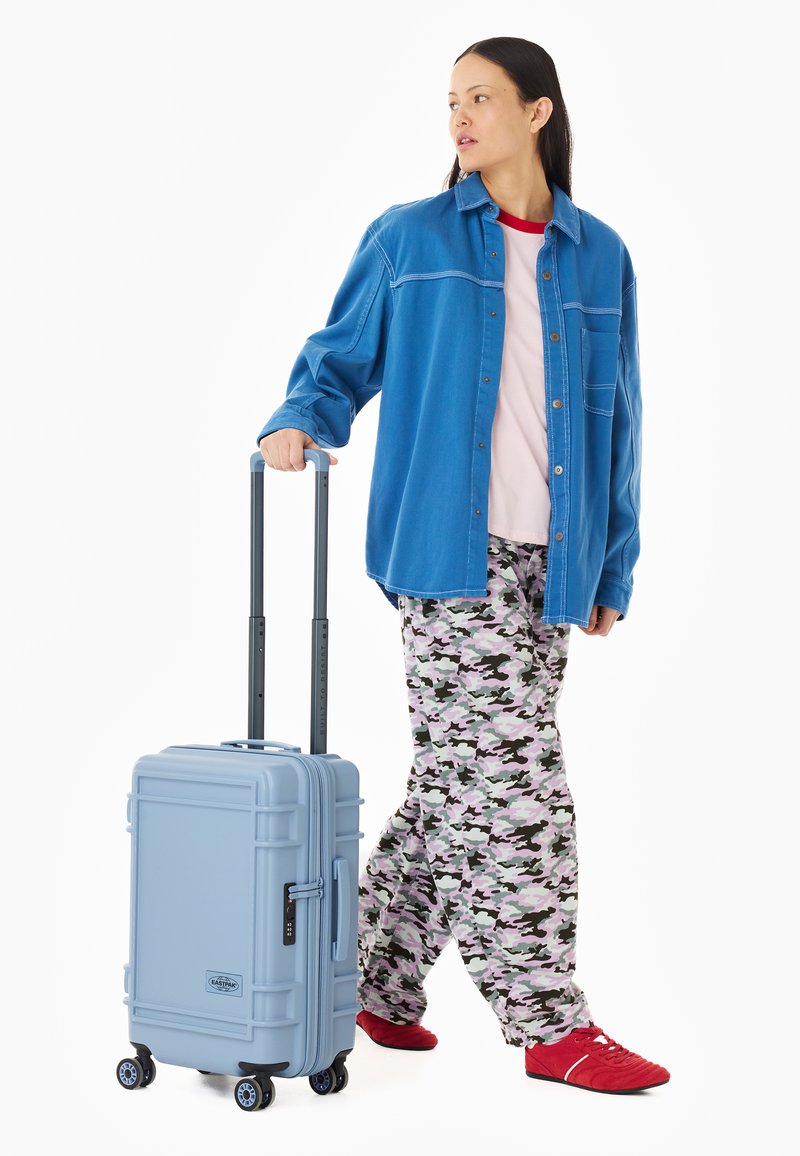 Jeune personne portant une veste bleue, un pantalon à motifs et des chaussures rouges, tenant la poignée d'une valise roulante bleu clair.