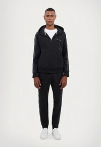 Melna džemperi ar rāvējslēdzēju un baltām svītrām gar piedurknēm, kopā ar atbilstošām sporta biksēm. Valkāts ar baltu T-kreklu un baltām sporta kurpēm.