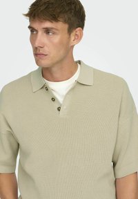 Sweter polo w jasnym beżu z krótkimi rękawami, płaskim kołnierzem i zapięciem na trzy guziki. Posiada teksturę ribbed i luźny krój.