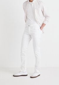 Białe dżinsy o slim fit, zestawione z jasną, pasiastą koszulą na guziki oraz białymi trampkami z szarymi akcentami. Gładka tekstura materiału.