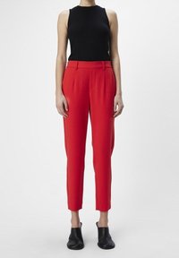 Pantalons ajustés rouges avec une coupe slim et deux poches avant, associés à un top sans manches noir. Le modèle porte des chaussures noires.