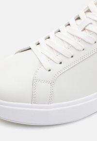 Sneaker in pelle bianca con lacci bianchi piatti, superficie testurizzata e suola in gomma liscia. Presenta cuciture e un design minimalista.