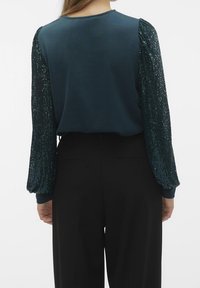 Top teal con maniche lunghe decorate con paillettes verdi, schiena liscia, scollo rotondo, abbinato a pantaloni neri.
