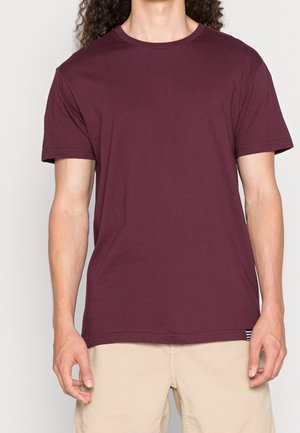 Person iført en ensfarvet kortærmet mørkerød t-shirt og beige shorts, stående mod en lys baggrund med armene afslappet ned langs siderne.