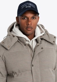 Gevoerde geblokte jas in kaki- en bruine tinten met een fleece-voering in de capuchon. Gecombineerd met een lichtgrijze hoodie en een marineblauwe pet met "NEW YORK" erop.