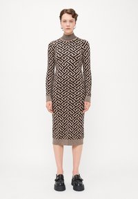 Strikket kjole med lange ermer som har et geometrisk mønster i brunt og beige, høy hals og ribbestrikkede mansjetter, iført chunky svarte sko.