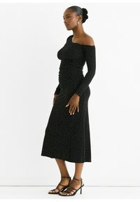 Black off-shoulder dress with long sleeves, ruched detailing, and white polka dots. Worn with strappy black high-heeled sandals.