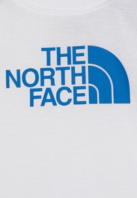 Biela bavlnená tričko s výrazným modrým logom s textom "THE NORTH FACE" a zakriveným dizajnovým prvkom vpravo.