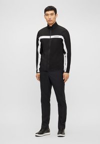 Veste zippée noire avec des rayures blanches sur les manches, assortie à un pantalon noir et des baskets noires. Tissu lisse avec une coupe ajustée.