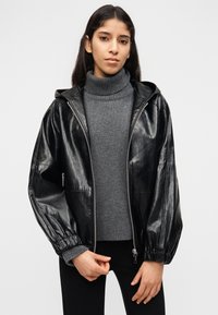 Νεαρή γυναίκα που φοράει μια γυαλιστερή μαύρη ζακέτα με φερμουάρ πάνω από γκρι πουλόβερ με ζιβάγκο και μαύρο παντελόνι, στέκεται μπροστά σε ένα απλό ανοιχτό φόντο.