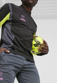 Zwart en grijs sportshirt met rits en gele accenten, gecombineerd met grijze broek. Het model houdt een felgele soccerbal vast.