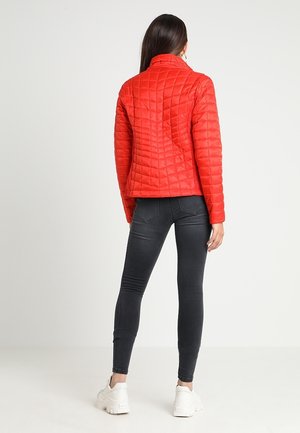 Nainen, jolla on kirkkaanpunainen tikkitakki, mustat kapeat farkut ja valkoiset lenkkarit, seisoo selin vaaleaa, yksinkertaista taustaa vasten.