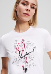 White t-shirt featuring black and red fashion sketches and handwritten notes on the front. Smooth texture with a classic crew neckline.