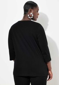 Top nero a maniche lunghe con una vestibilità ampia, scollo rotondo e una texture morbida. Presenta una leggera caduta sul retro ed è abbinato a grandi orecchini a cerchio in argento.