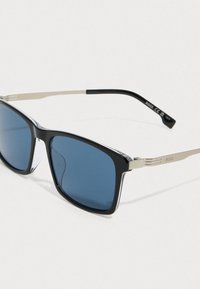 Melnas un sudraba saulesbrilles ar zilajiem tonētiem stikliem un plāniem metāla rokturīšiem, uz viena rokturīša iegravēts zīmola nosaukums "BOSS."