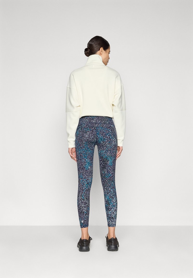 Weißes Sweatshirt mit hohem Kragen, kombiniert mit knöchellangen, mehrfarbigen Leggings mit einem strukturierten, abstrakten Muster in Blau, Schwarz und Weiß.