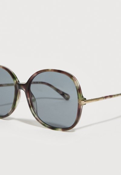 Υπερμεγέθη γυαλιά ηλίου με μοτίβο χελώνας, στρογγυλούς γκρι φακούς και λεπτούς χρυσούς μεταλλικούς βραχίονες, τοποθετημένα πάνω σε λευκή επιφάνεια.