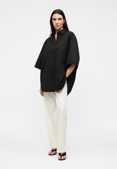 Γυναίκα που στέκεται με τα χέρια στις τσέπες, φοράει υπερμεγέθη μαύρο πουκάμισο με κουμπιά, λευκό παντελόνι με φαρδιά μπατζάκια και μαύρα σανδάλια σε λευκό φόντο.