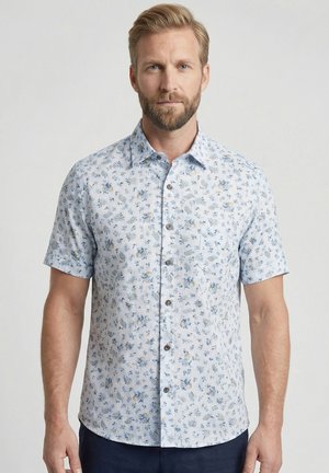 Man met baard die een wit overhemd met korte mouwen draagt met een klein blauw en beige bloemmotief, staand tegen een effen lichte achtergrond.