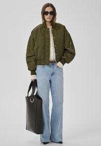 Frau trägt übergroße olivgrüne Bomberjacke, cremefarbenen Pullover, hellblaue Jeans mit weitem Bein, schwarze spitze Schuhe, Sonnenbrille, hält große schwarze Tragetasche.