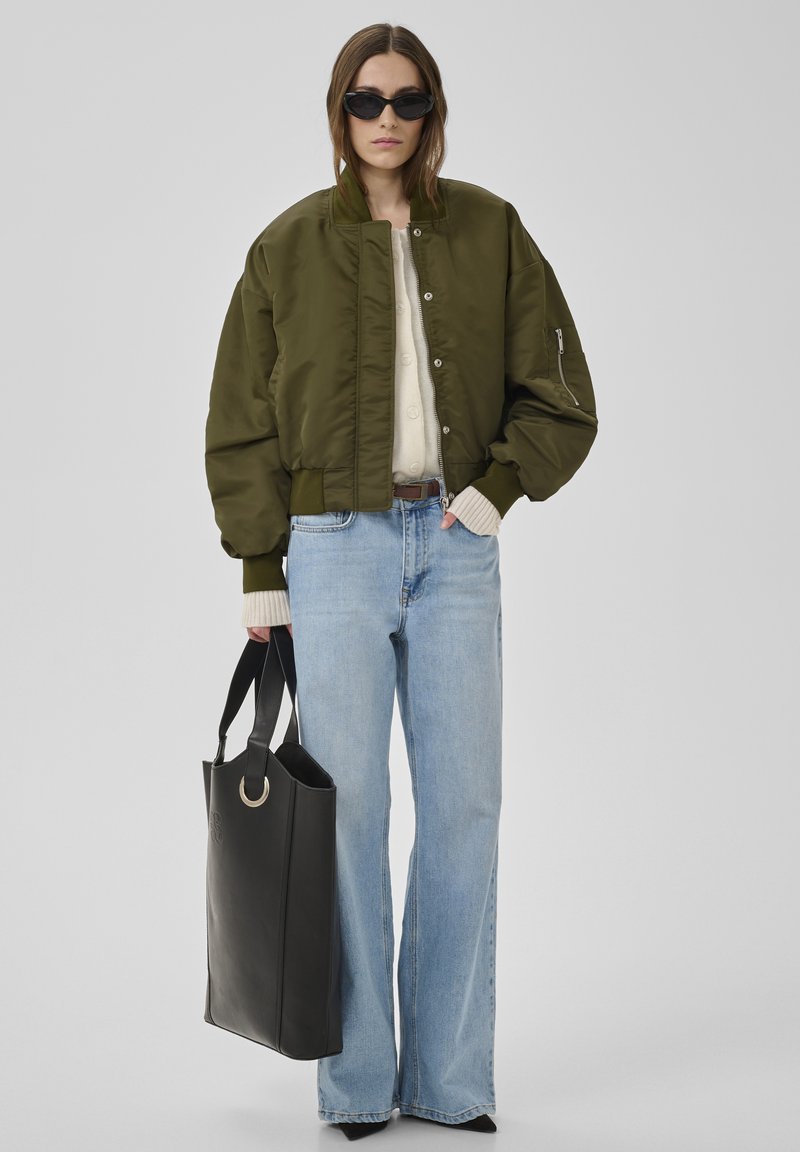 Frau trägt übergroße olivgrüne Bomberjacke, cremefarbenen Pullover, hellblaue Jeans mit weitem Bein, schwarze spitze Schuhe, Sonnenbrille, hält große schwarze Tragetasche.