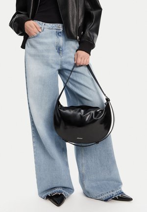 Persona que lleva vaqueros anchos azul claro, zapatos negros puntiagudos y chaqueta de cuero negra, sosteniendo un bolso negro brillante en forma de media luna.