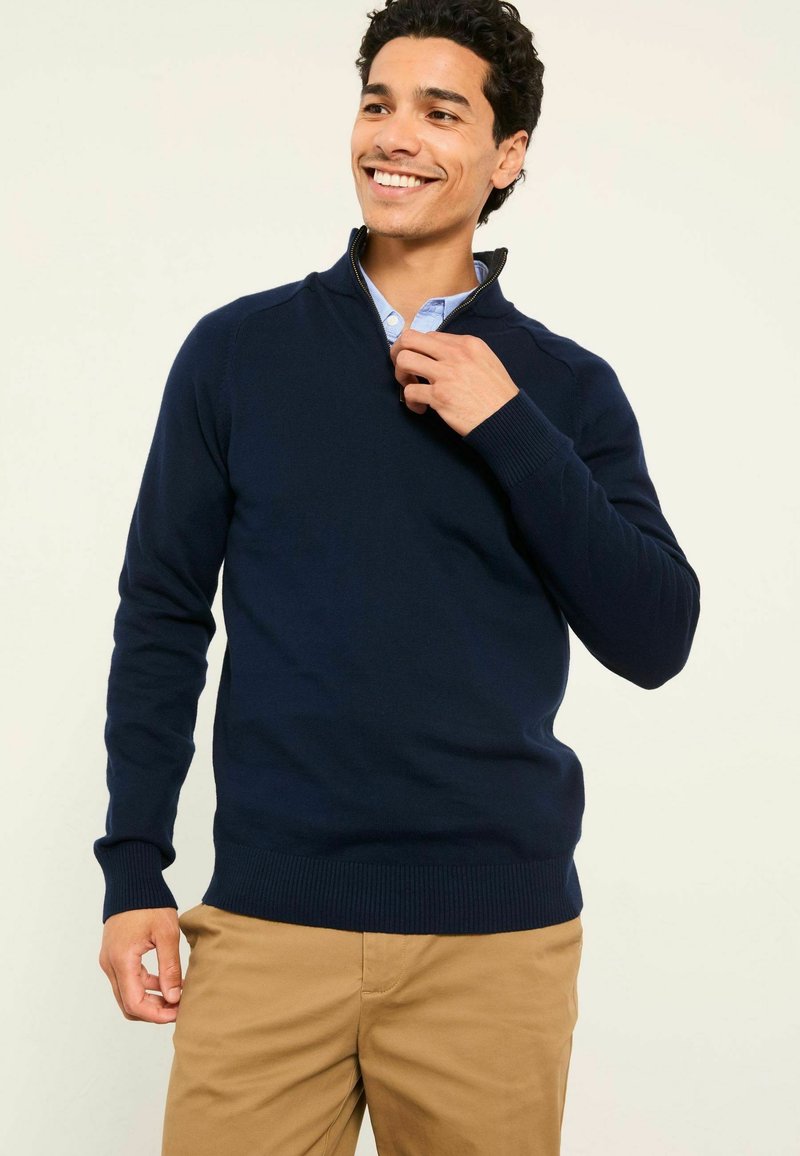 Dunkelblauer Pullover mit halbem Reißverschluss, gerippten Bündchen und Saum. Trägt man über einem hellblauen Hemd mit Kragen, aus Strickstoff.