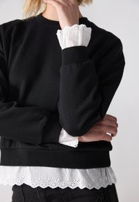Sweat-shirt noir avec poignets côtelés, porté par-dessus un chemisier blanc orné d'une bordure en dentelle ajourée festonnée le long de l'ourlet et des manches.