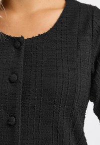 Cardigan nero in tessuto testurizzato con scollatura a girocollo, tre bottoni rotondi e un sottile motivo a griglia nel tessuto.