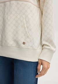 Hoodie crème avec un motif texturé, doté d'une poche frontale et de poignets côtelés. Détail de logo subtil en bronze sur le bas.