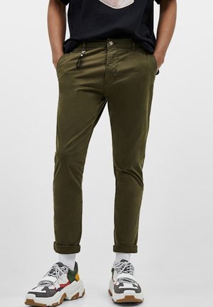 Unterkörper einer Person, die olivgrüne hochgekrempelte Hose, mehrfarbige klobige Sneaker, weiße Socken und ein schwarzes Shirt mit Händen in den Taschen trägt.