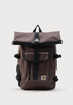 Mochila marrón con cierre enrollable, correa con hebilla negra, bolsillo frontal con cremallera, bolsillo lateral de malla y parche con el logo de Carhartt en la parte inferior derecha.