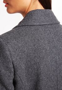 Blazer grigio in lana con una texture morbida, caratterizzato da un ampio colletto e una silhouette sagomata. Cuciture visibili lungo il bordo del colletto.