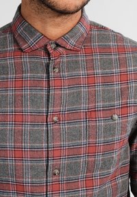 Camicia a quadri con colori rosso e grigio, con dettagli blu. Design con colletto, chiusura a bottoni e texture morbida. Due tasche sul petto con bottone.