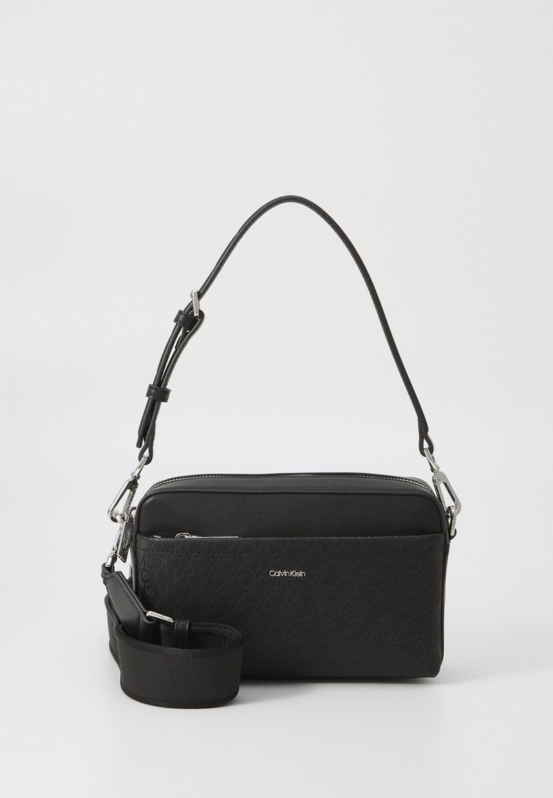 Czarna prostokątna torba z teksturowanym przodem, srebrnymi elementami metalowymi i odpinanym paskiem. Posiada zamek błyskawiczny oraz logo z przodu.