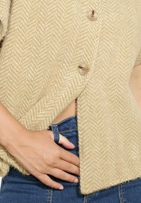 Cardigan in maglia a spina di pesce beige con una texture morbida e due bottoni trasparenti. Presenta una vestibilità ampia e spacco laterale, abbinato a jeans denim blu.