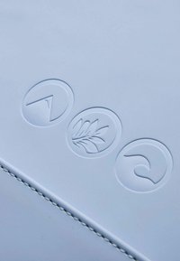 Weiße Oberfläche mit drei geprägten kreisförmigen Logos: einem Gipfel, einem Blatt und einer Welle, jeweils mit unterschiedlichen Designs. Genähte Kanten sind sichtbar.