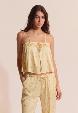 Frau trägt gelbes ärmelloses Oberteil mit Blumenmuster und dünnen Trägern sowie passende lockere Hose mit Kordelzug, steht vor beigem Hintergrund.