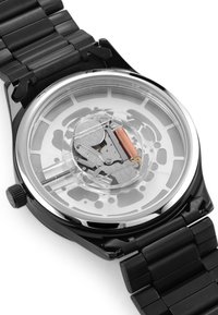 Svart rustfritt stålklokke med en gjennomsiktig bakside som viser de indre mekanismene, glatte lenker og en polert sølvbezel.