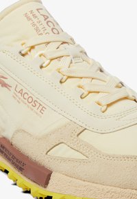 Zapatillas Lacoste en amarillo claro con acentos de ante; presentan una suela de goma texturizada, un logo en relieve y cordones planos.